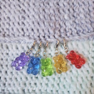 May include: Five colourful gummy bear stitch markers in purple, blue, green, yellow and red. The markers are attached to silver metal rings.