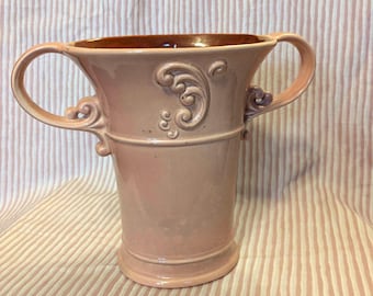 May include: A pale pink ceramic vase with two handles and a decorative design. The vase has a brown rim and a glossy finish.