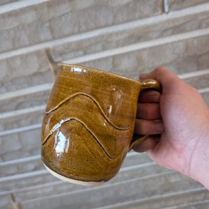 May include: A handmade ceramic mug with a brown glaze. The mug has a wavy design around the body and a rounded handle. The mug is held in a hand.