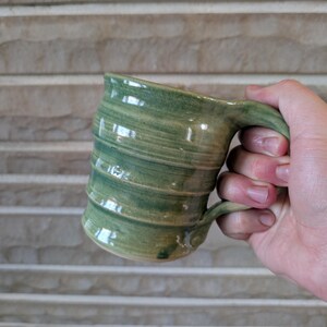 May include: A handmade ceramic mug with a green and tan swirl pattern. The mug has a curved handle and a slightly flared rim. The mug is held in a hand, showcasing its size and shape. The mug's exterior has a textured, organic feel.