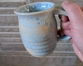 May include: Handmade ceramic mug with a light blue and tan glaze. The mug features a curved handle and a textured, ridged design around the body. The interior and exterior have speckled blue accents, creating a unique, artisanal look.