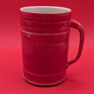 May include: A tall, red ceramic mug with a handle. The mug features a textured design around the middle and near the top and bottom. The interior is white. The mug is set against a red background.