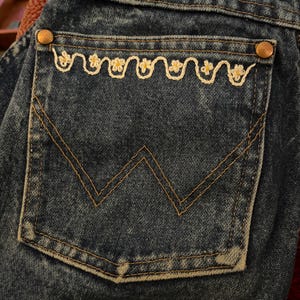 Könnte beinhalten: Nahaufnahme einer blauen Jeans-Gesäßtasche mit einem weißen, gestickten Gänseblümchen-Kettenmuster. Die Tasche hat braune Nähte und einen kleinen Metallknopf.