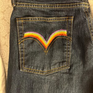 May include: Dark blue denim jeans with a back pocket featuring a rainbow-colored embroidered arc.