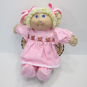 May include: A vintage doll with blonde yarn hair, blue eyes, and a soft, round face. The doll wears a pink and white gingham dress and matching pants, with teddy bear embroidery. Pink and white striped bows adorn the doll's hair.
