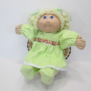 May include: A vintage doll with blonde yarn hair, blue eyes, and a soft, fabric face. The doll wears a light green checkered dress and pants with white lace trim. The doll is seated in a small woven basket.