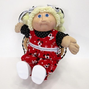May include: A vintage doll with blonde curly hair, blue eyes, and a round face. The doll wears a red outfit with a white dog pattern and black polka dot sleeves. It is seated in a woven basket chair.
