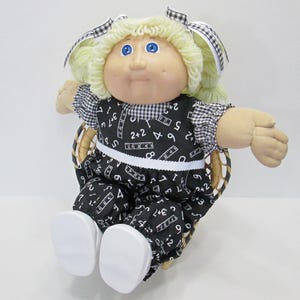 May include: A vintage doll with blonde yarn hair, blue eyes, and a round face. The doll wears a black and white patterned outfit with numbers and rulers, and white shoes. It is seated in a wicker chair.