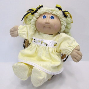 May include: A Cabbage Patch Kid doll wearing a yellow and white striped dress with a bee and flower design. The doll has blonde yarn hair with black and yellow bows. The doll is sitting in a wicker chair.