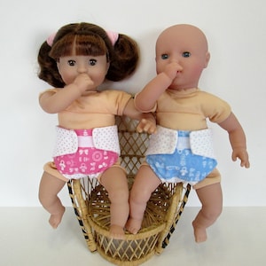 May include: Two baby dolls with fair skin and light brown hair, wearing peach-colored shirts and patterned diapers. One doll has pink diapers, the other blue. They are seated in a small wicker chair.