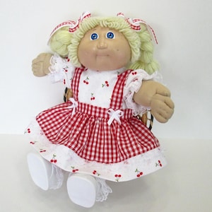 May include: A vintage doll with blonde yarn hair styled in pigtails, adorned with red and white ribbons. The doll wears a red and white checkered dress with cherry accents and white lace trim. The doll has blue eyes and a soft, fabric body.
