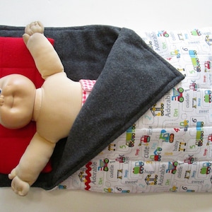 May include: A gray and white quilted sleeping bag with a red pillow inside. A doll is lying in the sleeping bag with its arms outstretched. The sleeping bag has a pattern of construction vehicles and the words "Adorable", "Dirt", "Cuddle", "Beep", "Sweet", "Hugs", "Stop", "Cute", and "Baby" on it.