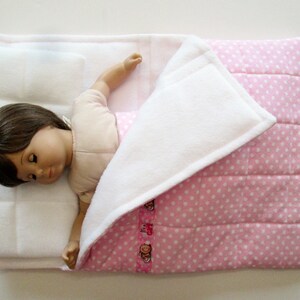 May include: A white and pink doll bed with a white pillow and a doll sleeping in it. The doll is wearing a pink and white dress.