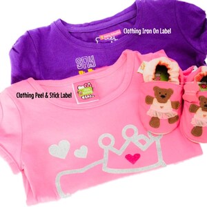 May include: Two pink and purple t-shirts with a crown and heart design on the pink shirt. The pink shirt has a peel and stick label that says "Say Hello" and the purple shirt has an iron-on label that says "Clothing Iron On Label". There are also two pink baby shoes with a brown bear design.