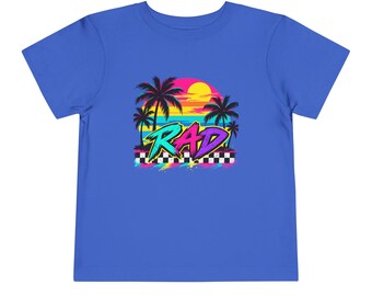 以下が含まれることがあります： ロイヤルブルーの半袖Tシャツ。ヤシの木があるビーチの夕日と、黒と白の市松模様の上にスタイリッシュなフォントで書かれた「RAD」という文字のグラフィックプリントが施されています。鮮やかなネオンカラーが使用されています。