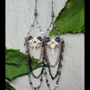 May include: A pair of dangle earrings featuring white bone vertebrae centers flanked by purple beads. The design includes layered chains in silver, copper, and black tones, suspended from silver-toned ear wires in a teardrop-shaped arrangement.