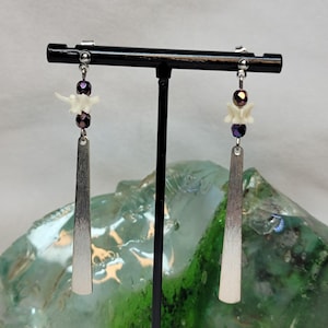 May include: A pair of dangle earrings featuring a small white bone vertebra segment, two dark metallic purple faceted beads, and a long, flat, brushed silver-tone metal bar pendant suspended from a silver-tone post and earring back.