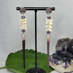 May include: A pair of dangle earrings featuring a silver-toned hammered disc at the top, followed by a purple faceted bead, a small white bone segment, another purple bead, and a long, slender, dark brown, pointed spike-shaped pendant at the base.
