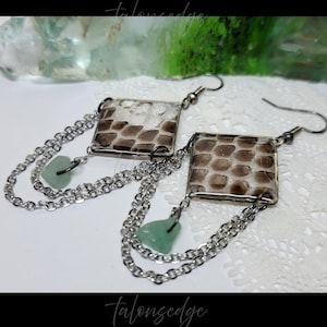 May include: A pair of dangle earrings featuring square frames with a brown and white snakeskin pattern. Each earring has two silver-toned metal chains draped below the square, accented with a small, pale green, irregular-shaped stone bead.
