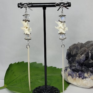 May include: A pair of dangle earrings featuring silver-tone metal bows at the top, clear faceted crystal beads, small off-white animal vertebrae, and long, thin, pointed off-white bone spikes hanging from the bottom of each earring.