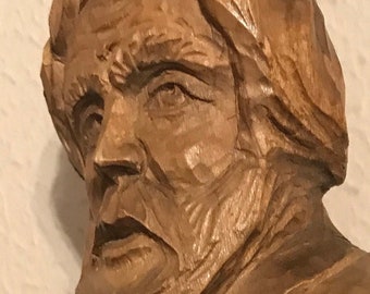 Op de afbeelding: Een gesneden houten sculptuur die een oudere man met een baard en een jongere persoon afbeeldt. De sculptuur heeft een warme, natuurlijke houttint, met zichtbare gesneden details. De figuren zijn gedeeltelijk gedrapeerd.