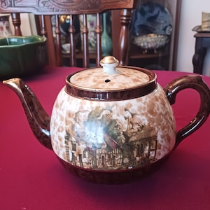 May include: A vintage ceramic teapot with a brown and cream speckled pattern. The teapot features a landscape scene and a dark brown handle and spout. The lid has a gold-colored knob. The teapot is sitting on a red surface.