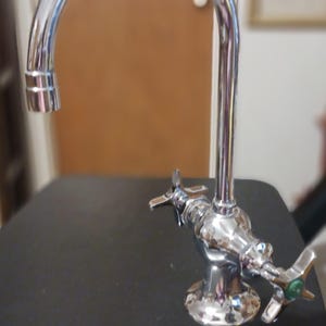 May include: A chrome-finished faucet with a high-arc spout and two cross-style handles. One handle features a green circular accent. A metal ruler is positioned at the base, showing the faucet width is approximately 6 inches across.