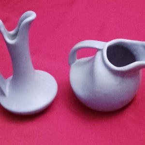 May include: Two light blue ceramic pitchers. One is tall with a curved handle and a flared top. The other is round with a handle and a spout. Both are on a red background.