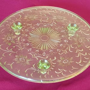 May include: A vintage, three-legged, clear glass cake stand with a delicate floral pattern. The stand is made of uranium glass, which glows a pale green under a blacklight.