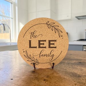 May include: A round, light brown cork trivet featuring a dark brown laser-engraved design. The text reads "the LEE family" in a combination of script and block lettering, framed by a circular border with decorative leaf sprigs on the sides.