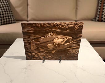May include: A wooden plaque featuring a detailed relief carving of a fish with an open mouth, set against a background of water and reeds. The artwork is in shades of brown and is displayed on a white table.