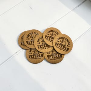 May include: A collection of circular light brown cork coasters featuring a dark brown laser-engraved design. Each coaster displays an eagle head icon above the text UNITED STATES VETERAN, accented with stars and horizontal lines on both sides.