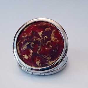 May include: A silver compact mirror with a red and gold swirl design. The mirror is closed.