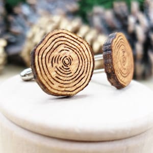 Puede incluir: Un par de gemelos de madera con un diseño de anillo de árbol. Los gemelos son de color marrón y tienen un acabado de madera natural. Se muestran en una caja blanca.
