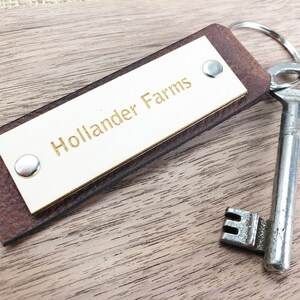以下が含まれることがあります： 「Hollander Farms」と刻まれた金色の金属タグが付いた茶色の革製のキーホルダー。シルバーの鍵がキーホルダーに取り付けられています。