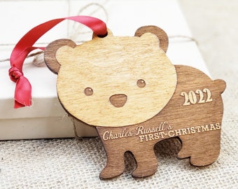 May include: Wooden bear Christmas ornament with a red ribbon. The ornament is light and dark brown, with the text "2022" and "Charles Russell's FIRST CHRISTMAS" engraved on it. The bear has a light brown face.