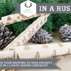 May include: Three wooden rolling pins with different engraved designs. The designs include a Christmas tree, reindeer, and a snowman. The rolling pins are on a burlap background with pine branches and pine cones.
