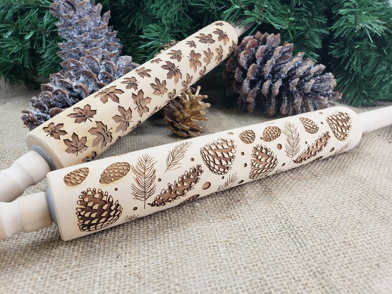 May include: Two wooden rolling pins with engraved designs. One pin features a pattern of maple leaves, while the other has a pattern of pine cones and branches.