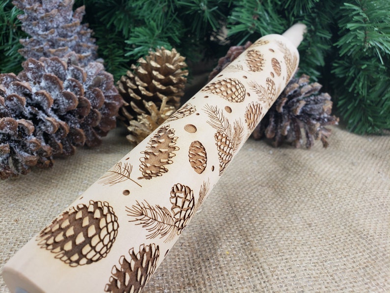May include: A wooden rolling pin with a design of pine cones and branches. The rolling pin is light brown and the design is dark brown.