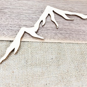 Op de afbeelding: Een houten uitsnijding van een bergketen met een natuurlijke houtkleur. De uitsnijding is op een jute achtergrond.