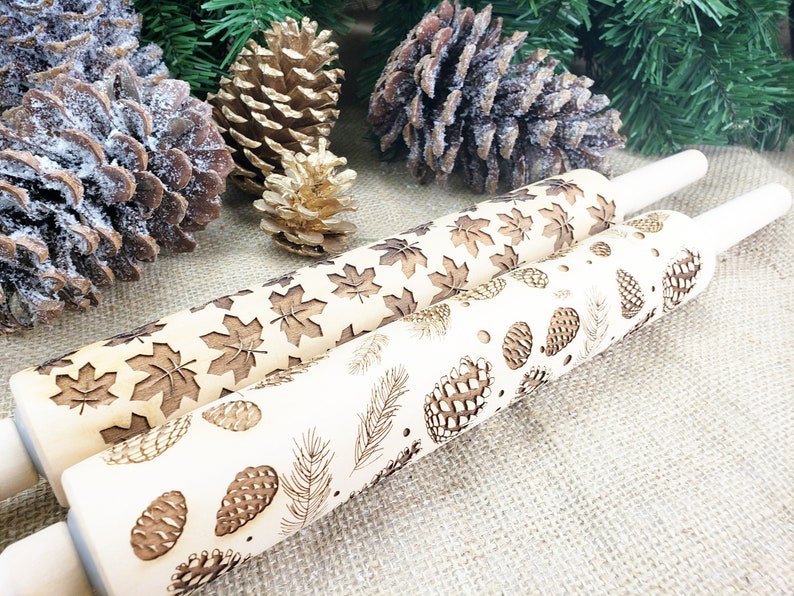 May include: Three wooden rolling pins with engraved designs of pine cones, leaves, and feathers. The rolling pins are on a hessian background.