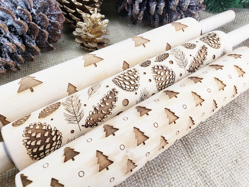 May include: Three wooden rolling pins with different Christmas-themed designs. The top rolling pin has a pattern of small Christmas trees. The middle rolling pin has a pattern of pine cones and feathers. The bottom rolling pin has a pattern of larger Christmas trees and small circles.