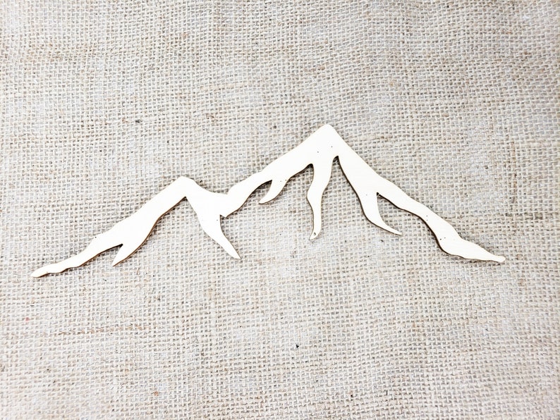 Op de afbeelding: Witte houten berg silhouet uitsnijding. De berg vorm is een eenvoudig ontwerp met een paar toppen en valleien. De uitsnijding is op een jute achtergrond.