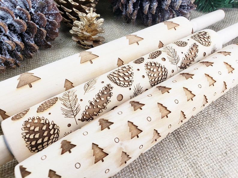 May include: Three wooden rolling pins with different Christmas-themed designs. The top rolling pin has a pattern of small Christmas trees. The middle rolling pin has a pattern of pine cones and feathers. The bottom rolling pin has a pattern of pine cones and small Christmas trees.