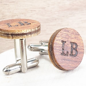 May include: A pair of wooden cufflinks with silver metal clasps. The cufflinks are round and have the initials "LB" engraved on them.