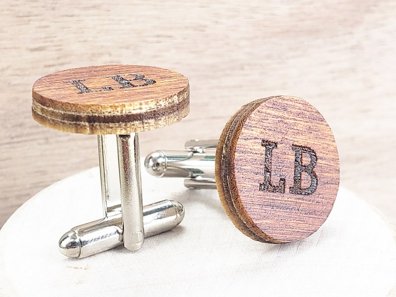 May include: A pair of wooden cufflinks with silver metal clasps. The cufflinks are round and have the initials "LB" engraved on them.
