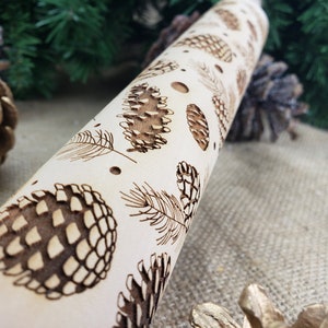 May include: A wooden rolling pin with a design of pine cones and branches. The rolling pin is light brown and the design is dark brown.