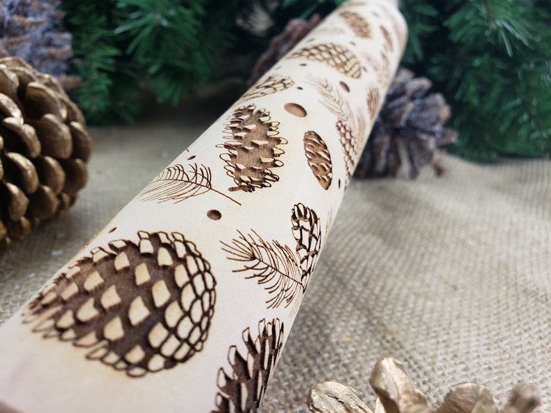May include: A wooden rolling pin with a design of pine cones and branches. The rolling pin is light brown and the design is dark brown.