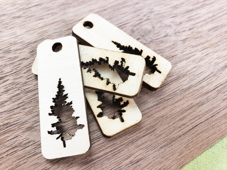 May include: Four wooden tags with a cutout design of a pine tree. The tags are light brown and have a hole at the top for hanging.