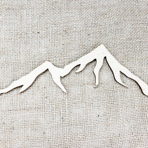 Op de afbeelding: Een wit houten berg silhouet uitsnijding, perfect voor knutselen of decoreren. De uitsnijding is op een jute achtergrond.